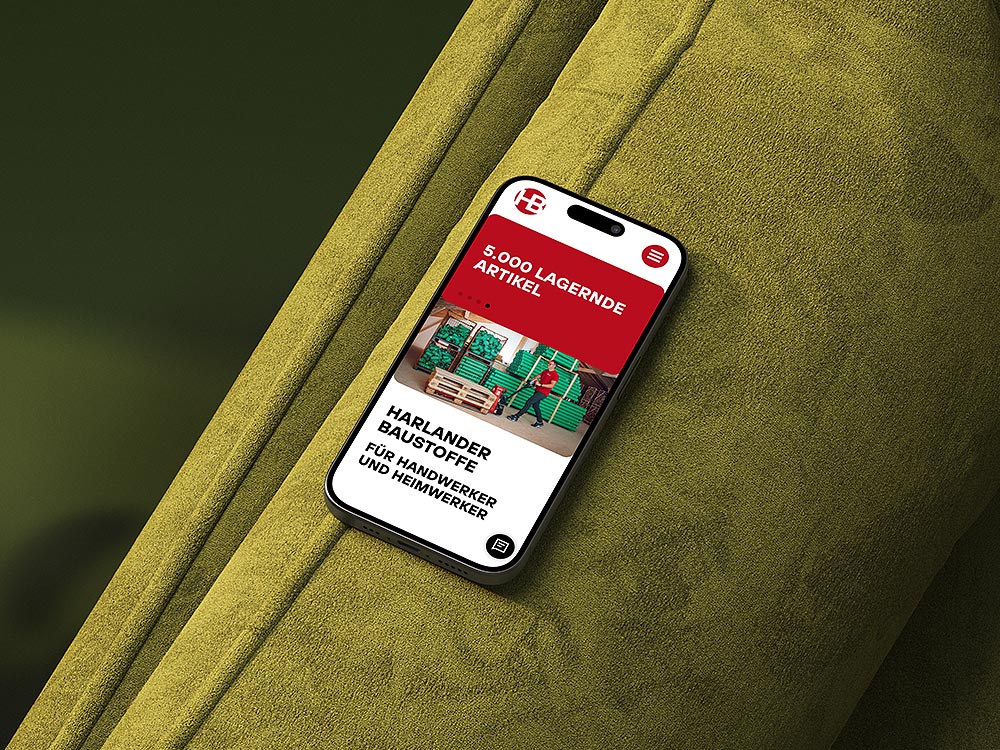 Smartphone mit geöffneter Website von Harlander Baustoffe, liegend auf einer grünen Stofffläche. Die Startseite zeigt den Text '5.000 lagernde Artikel – Harlander Baustoffe für Handwerker und Heimwerker' sowie das Bild eines Mitarbeiters, der einen Hubwagen bedient. Die Website ist für mobile Nutzung optimiert.