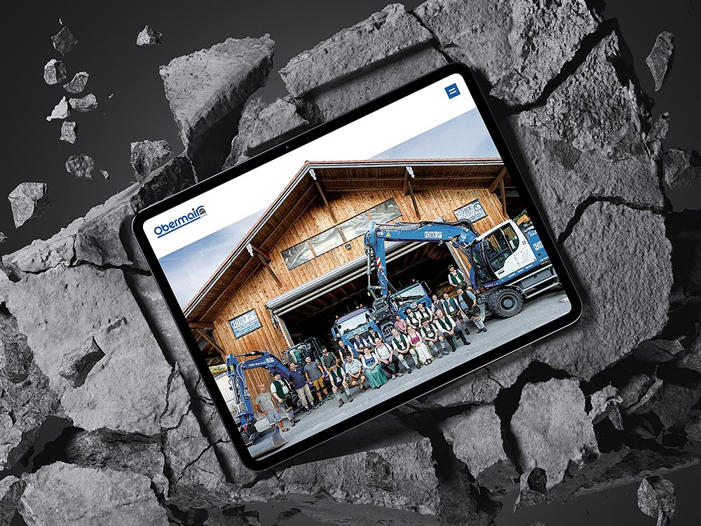 Tablet mit geöffneter Website von Obermair, platziert auf einer gebrochenen Betonfläche. Der Bildschirm zeigt ein Gruppenfoto des Bauunternehmens Obermair mit Team und Maschinen vor einem Firmengebäude. Die Website bietet ein modernes, SEO-optimiertes Webdesign.