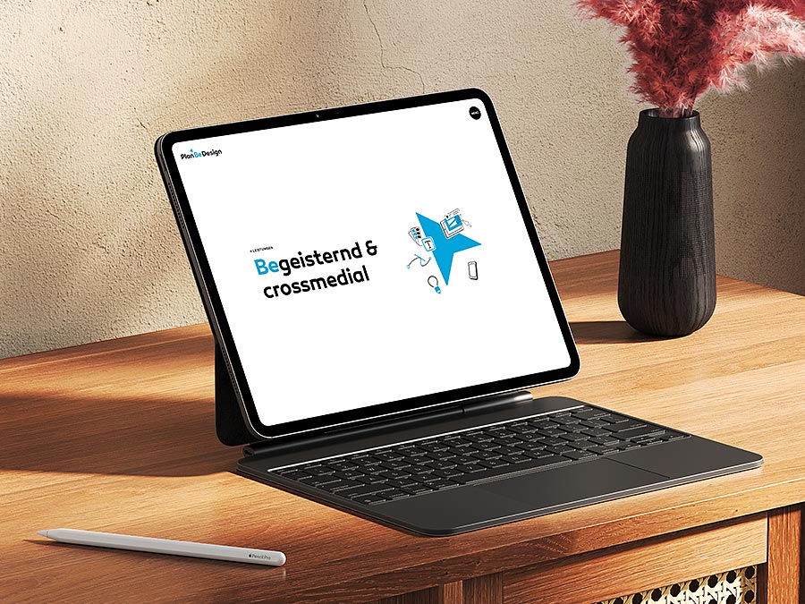 Tablet mit geöffneter Website von PlanBeDesign, der Kreativagentur hinter 'Love @ First Site'. Der Bildschirm zeigt den Slogan 'Begeisternd & crossmedial' mit einer blauen Grafik, die für innovative Crossmedia-Lösungen steht.