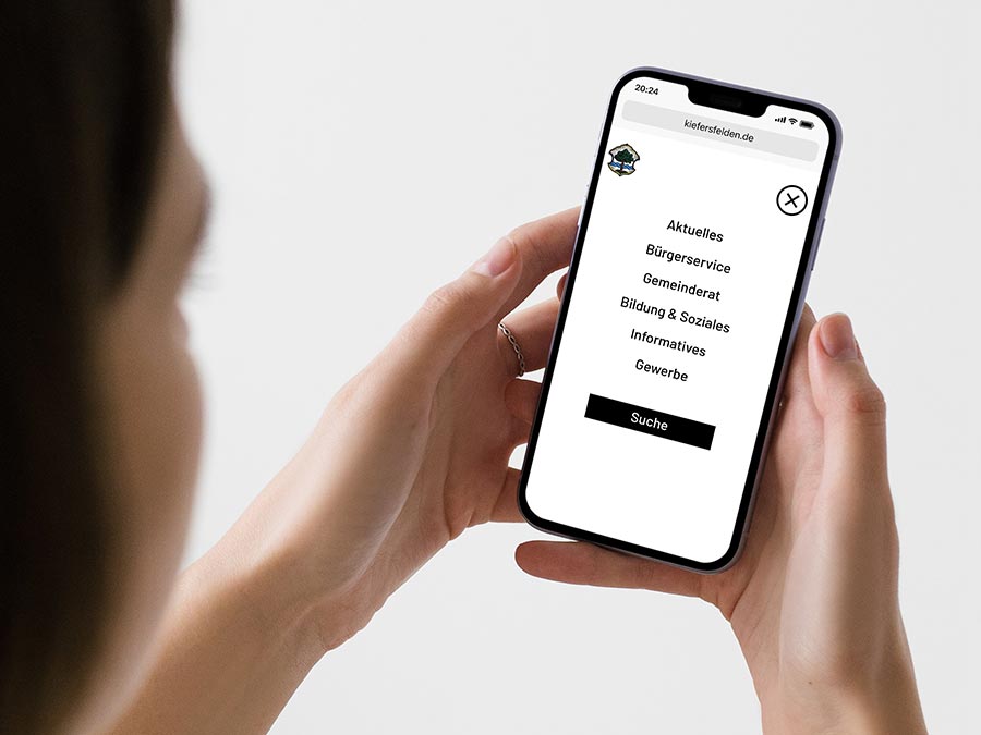 Smartphone mit geöffneter Website der Gemeinde Kiefersfelden, gehalten von einer Frau. Der Bildschirm zeigt ein strukturiertes Menü mit Kategorien wie 'Bürgerservice', 'Gewerbe' und 'Bildung & Soziales' für eine einfache Navigation.